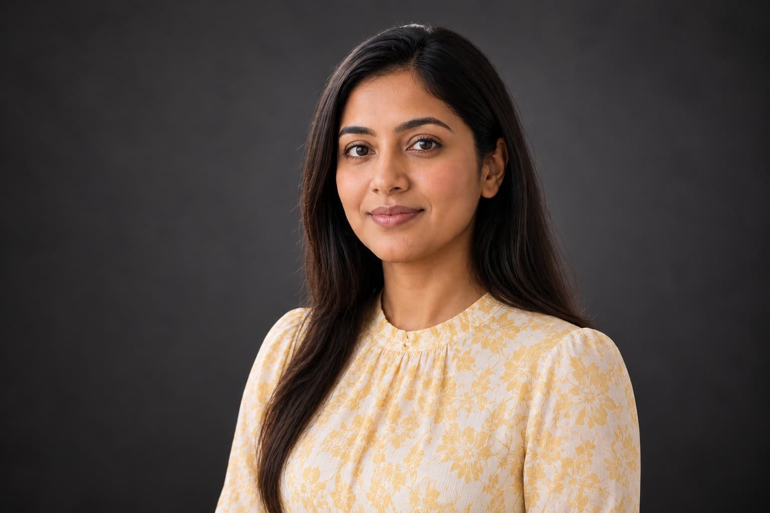 Smiling woman with long dark hair wearing a yellow floral blouse against a dark background.