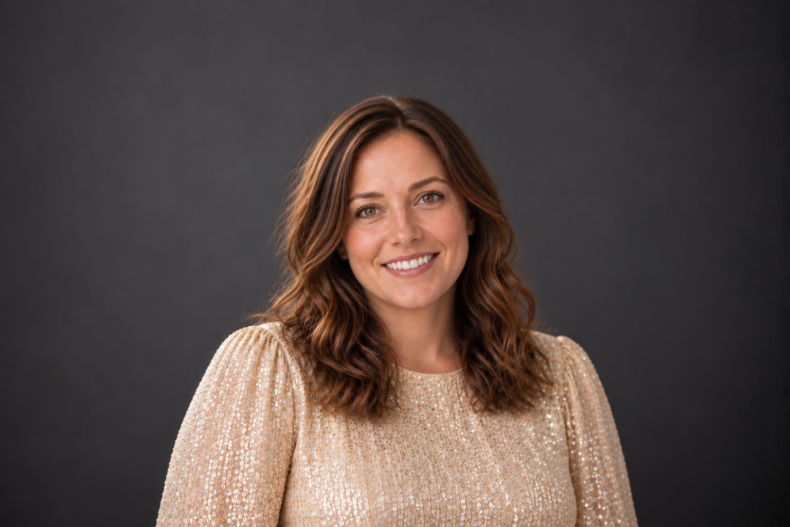 Smiling woman with wavy brown hair wearing a gold sequined top against a dark background.